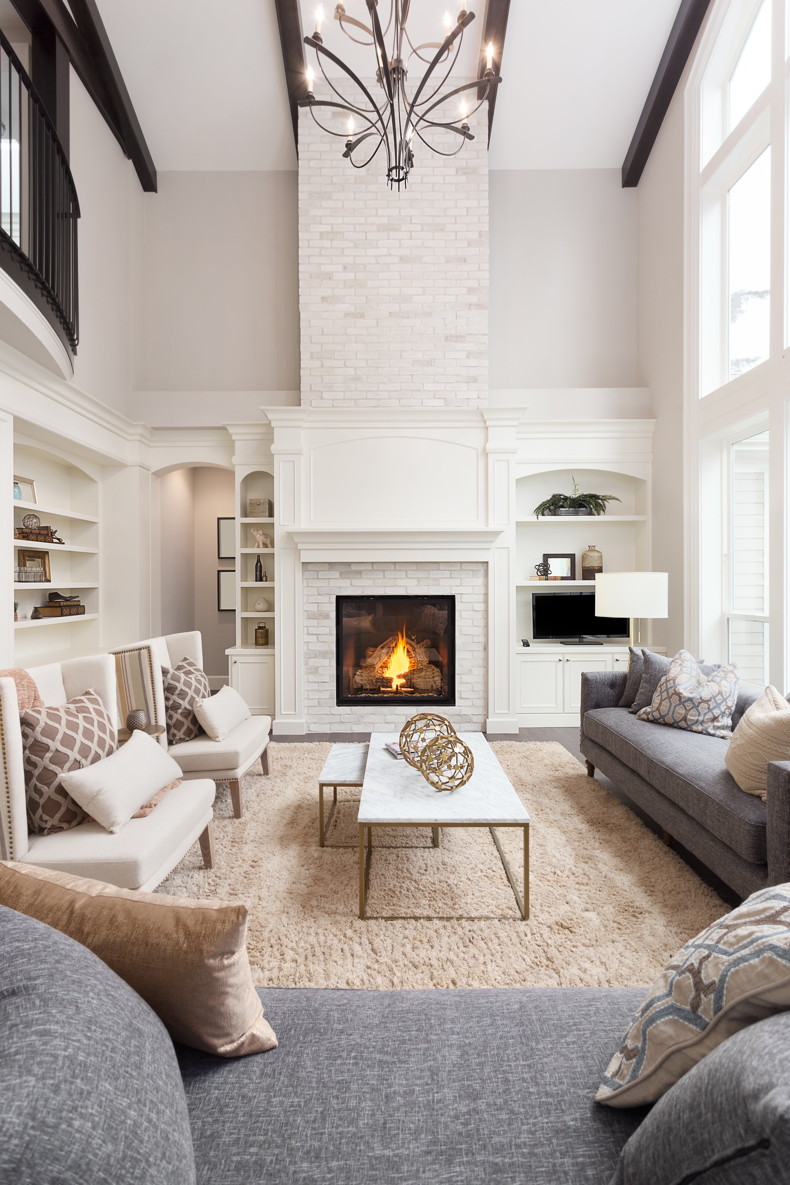 Bright, elegant living room with high ceilings, a lit fireplace, grey sofas, armchairs with patterned cushions, built‑in shelving, and a large chandelier overhead.
