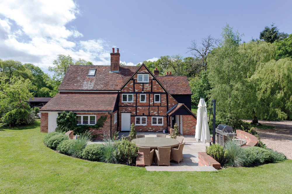 Exterior of a traditional brick cottage with a tiled roof, multiple small windows, and a well‑kept garden. In the foreground is a patio area with a round outdoor dining table, wicker chairs, potted plants, and a barbecue, surrounded by green lawn and trees under a bright sky.