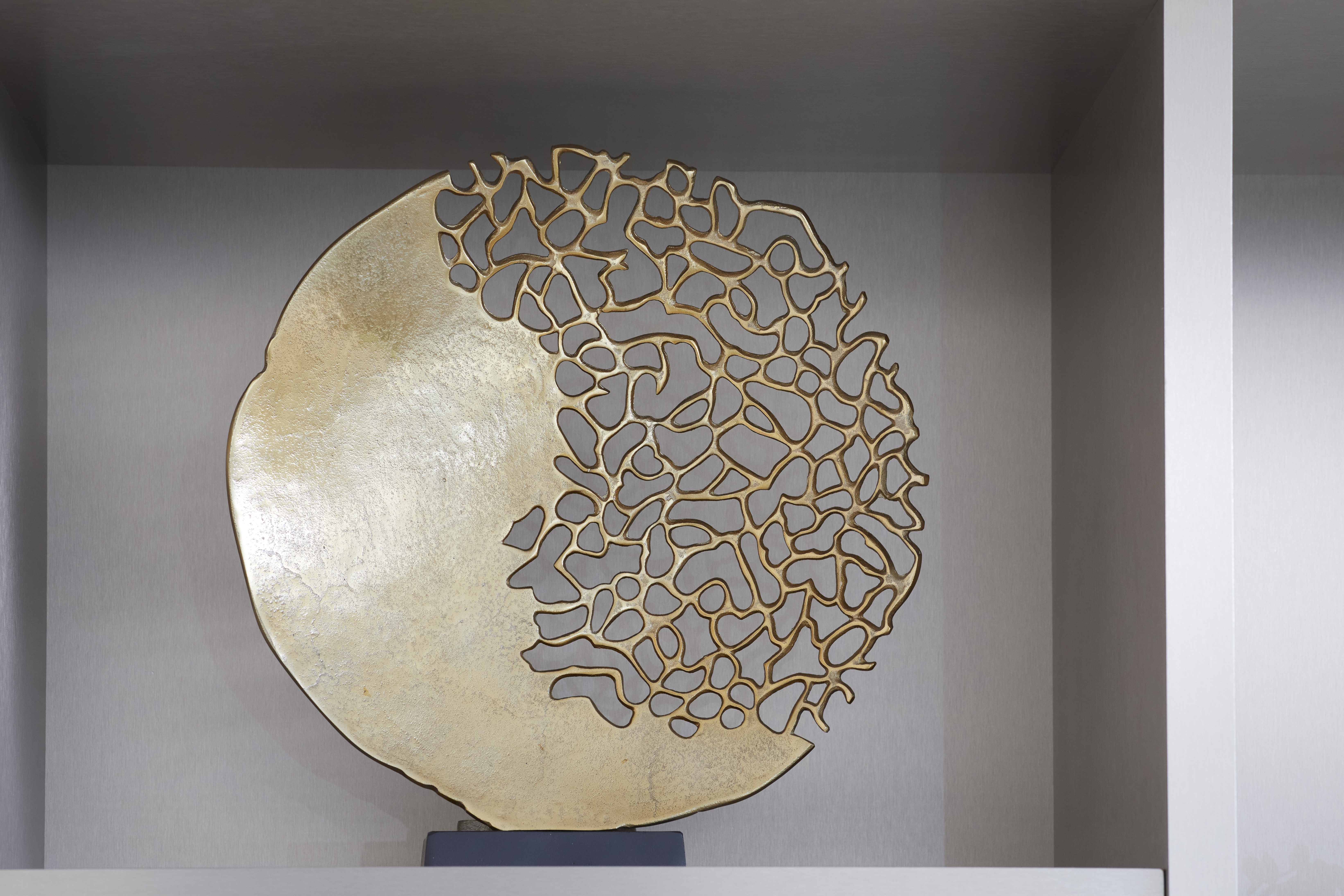 Decorative circular sculpture displayed on a shelf, with one half solid gold and the other half made of an open, irregular lattice pattern