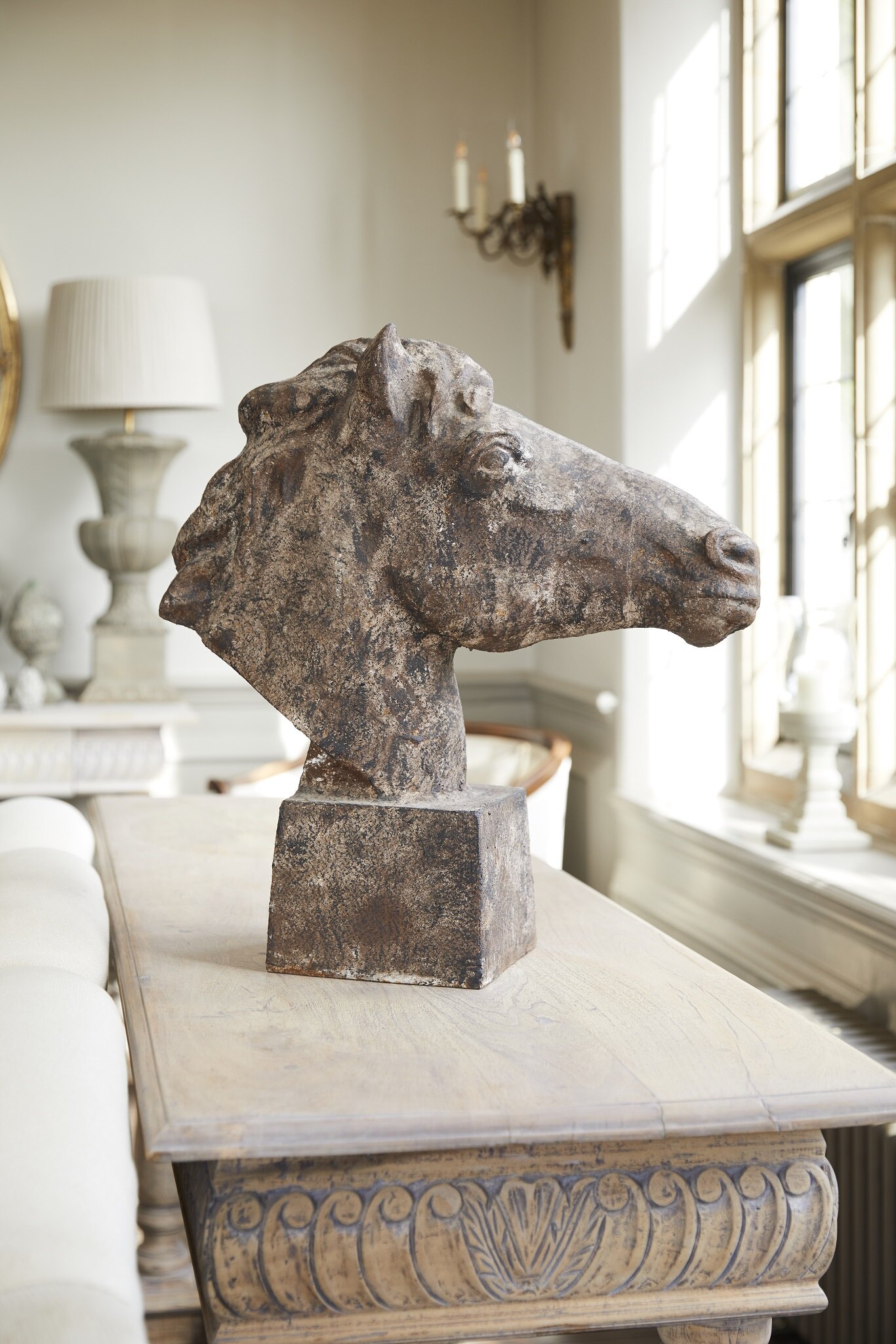 Stone sculpture of a horse’s head displayed on a carved wooden table in a traditional, light‑filled room with large windows and classic interior décor.