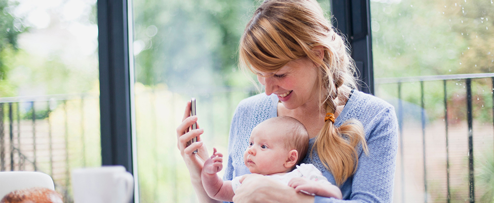 Auburn haired mother in blue cardigan showing young baby in white baby grow smartphone